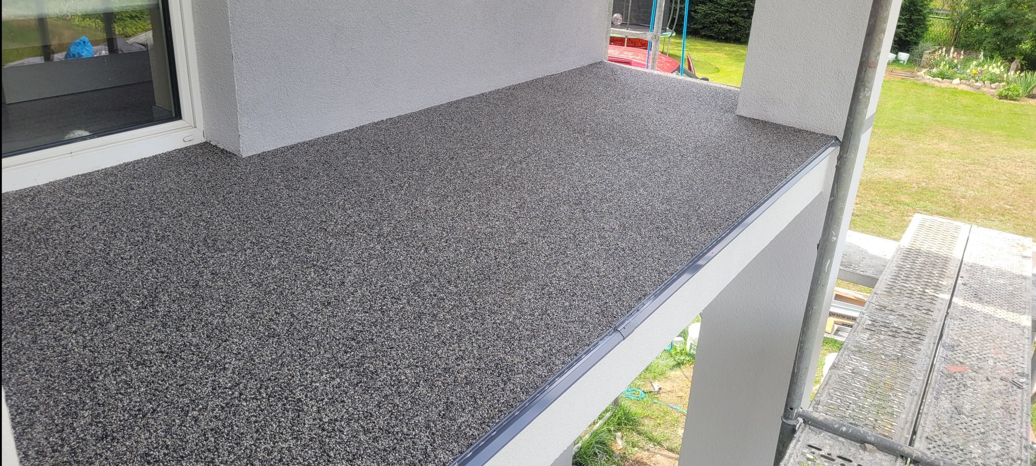 Nowoczesny taras z szarej, drobnoziarnistej nawierzchni żywicznej, wykończony listwą przypodłogową, widok z rusztowania na tle zielonego ogrodu.