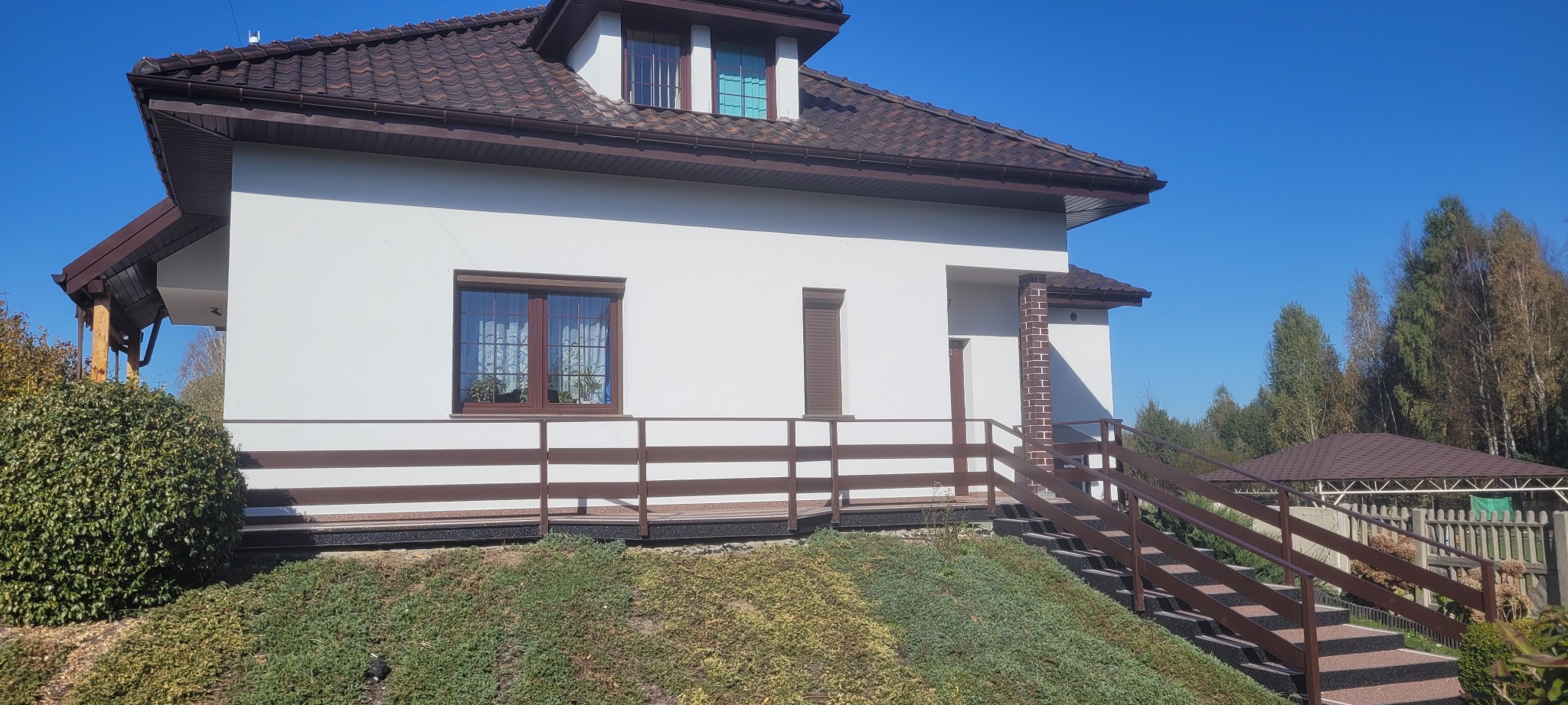 Ocieplony dom jednorodzinny z brązową stolarką okienną i schodami zewnętrznymi z barierkami, widok na ogród i błękitne niebo.