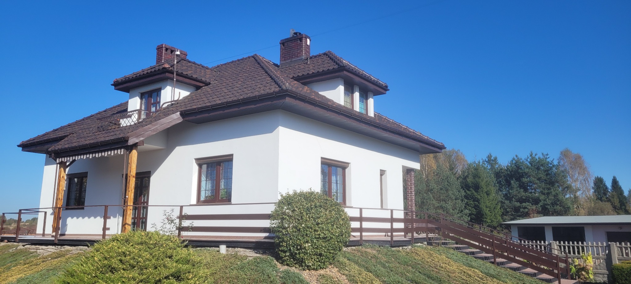 Biały dom jednorodzinny z brązowym dachem, drewnianymi elementami wykończeniowymi i schodami prowadzącymi do ogrodu, sfotografowany w słoneczny dzień.