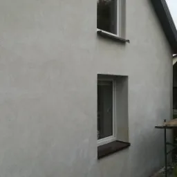 Elewacja domu jednorodzinnego z dwoma oknami po nałożeniu jasnoszarego tynku strukturalnego, widoczny fragment dachu i rusztowania.