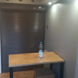 Szare wnętrze z drewnianym stołem, dwiema zielonymi krzesłami i zasłoniętymi żaluzjami.