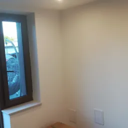 Świeżo pomalowane jasne ściany w narożnym pokoju z oknem i otwartymi drzwiami wejściowymi, widoczne skrzynki rewizyjne na ścianie.