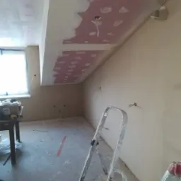 Poddasze w trakcie remontu: widoczne fragmenty sufitu z nałożoną masą szpachlową na łączeniach płyt, drabina aluminiowa, okno w tle, ślady pyłu na podłodze.