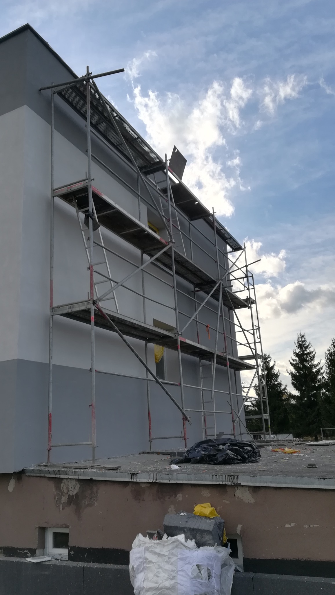 Świeżo pomalowana elewacja budynku w odcieniach szarości, z widocznym rusztowaniem na tle pochmurnego nieba. Na dachu budynku widoczne materiały budowlane i worki.
