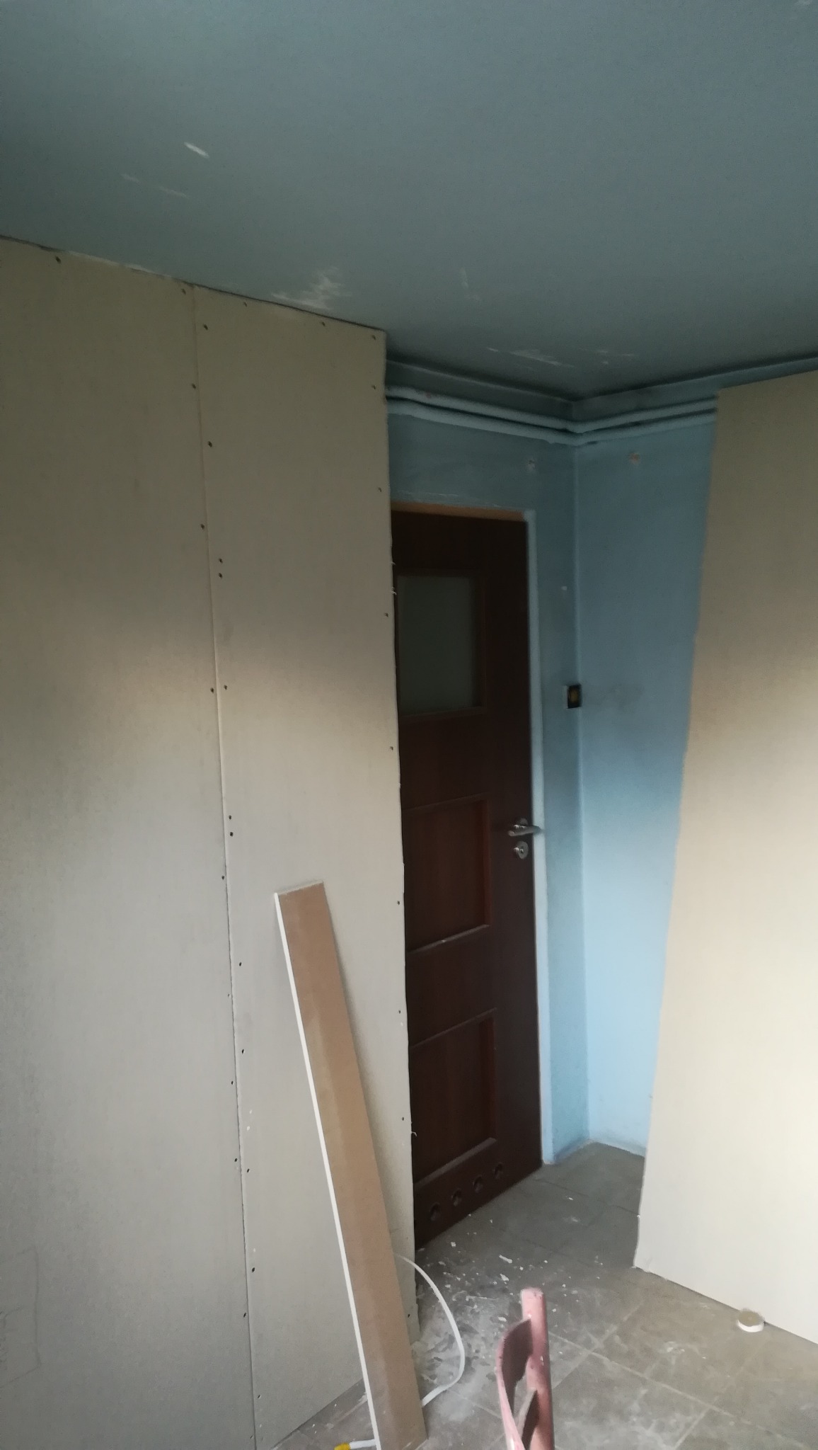 Montaż płyty karton gips na ścianie w trakcie remontu, widoczna drewniana listwa przypodłogowa i fragment brązowych drzwi.