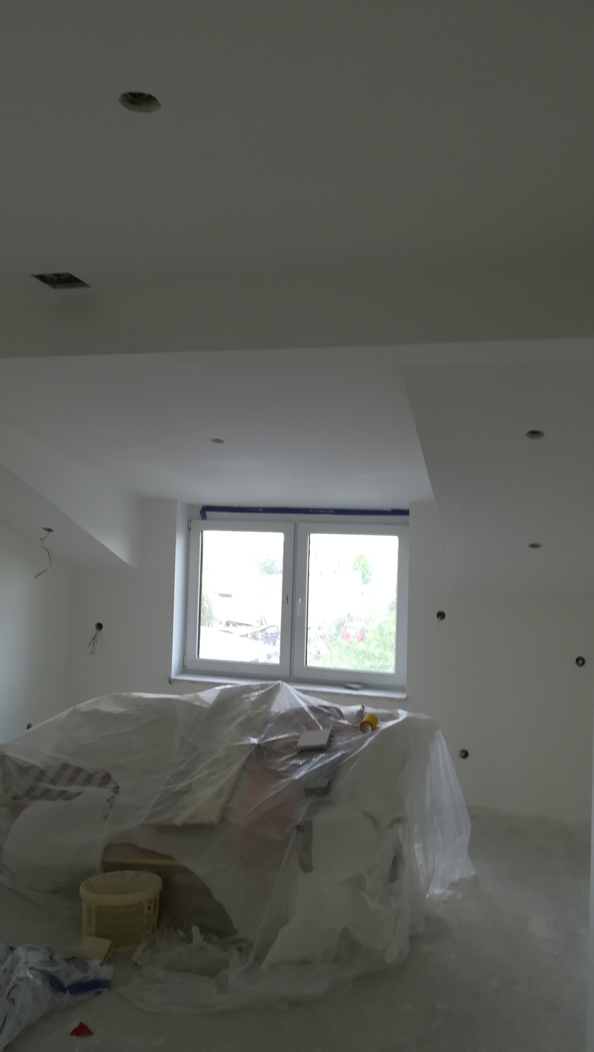 Wnętrze w trakcie remontu: świeżo pomalowane ściany i sufit na biało, okno zabezpieczone taśmą malarską, meble przykryte folią ochronną, widoczne puszki instalacyjne na ścianach i otwory...