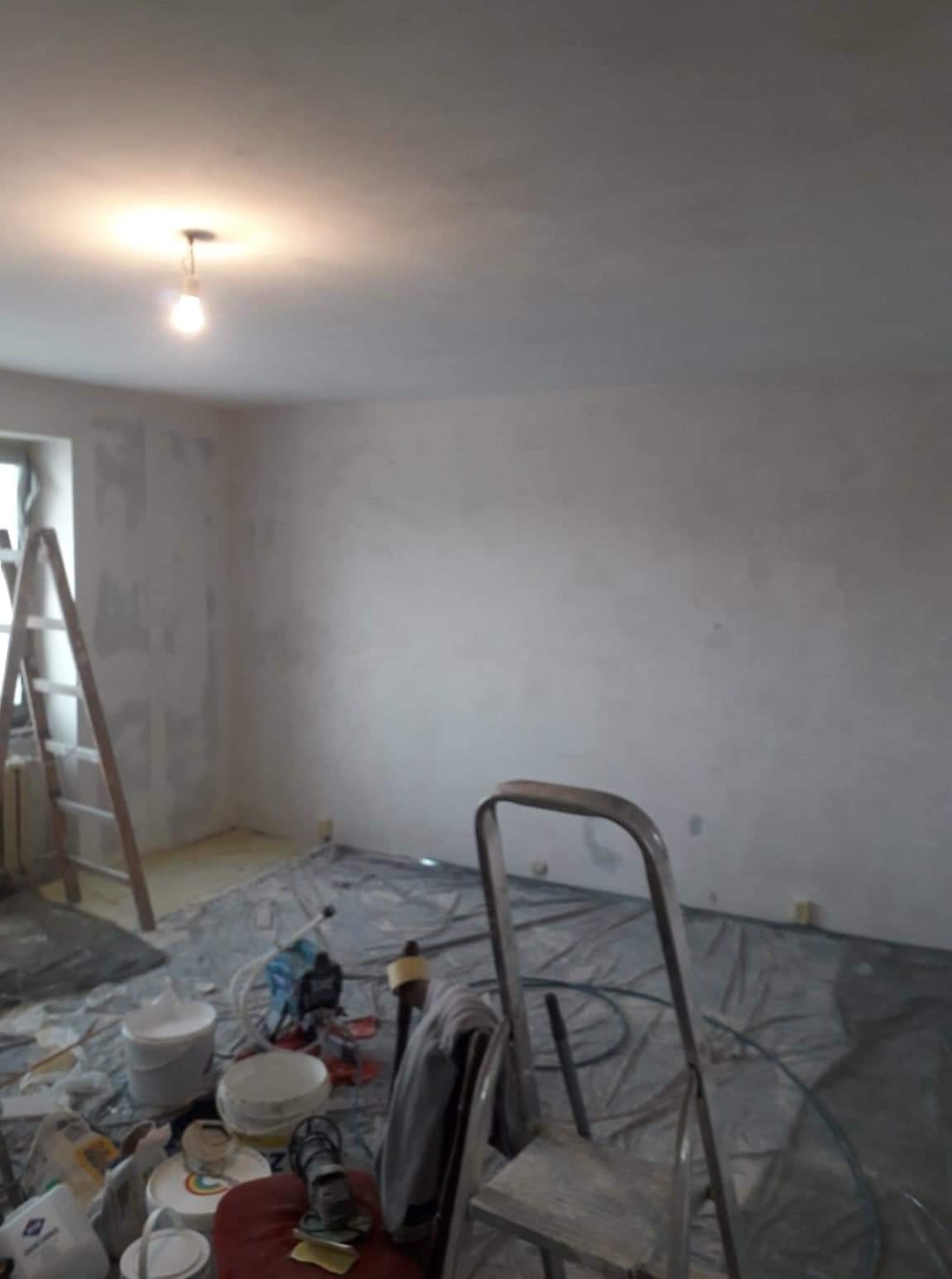 Pokój w trakcie remontu: ściany pokryte świeżo nałożoną warstwą, widoczne drabiny, wiadro z zaprawą i narzędzia malarskie na folii ochronnej rozłożonej na podłodze, sufit oświetlony pojedynczą...