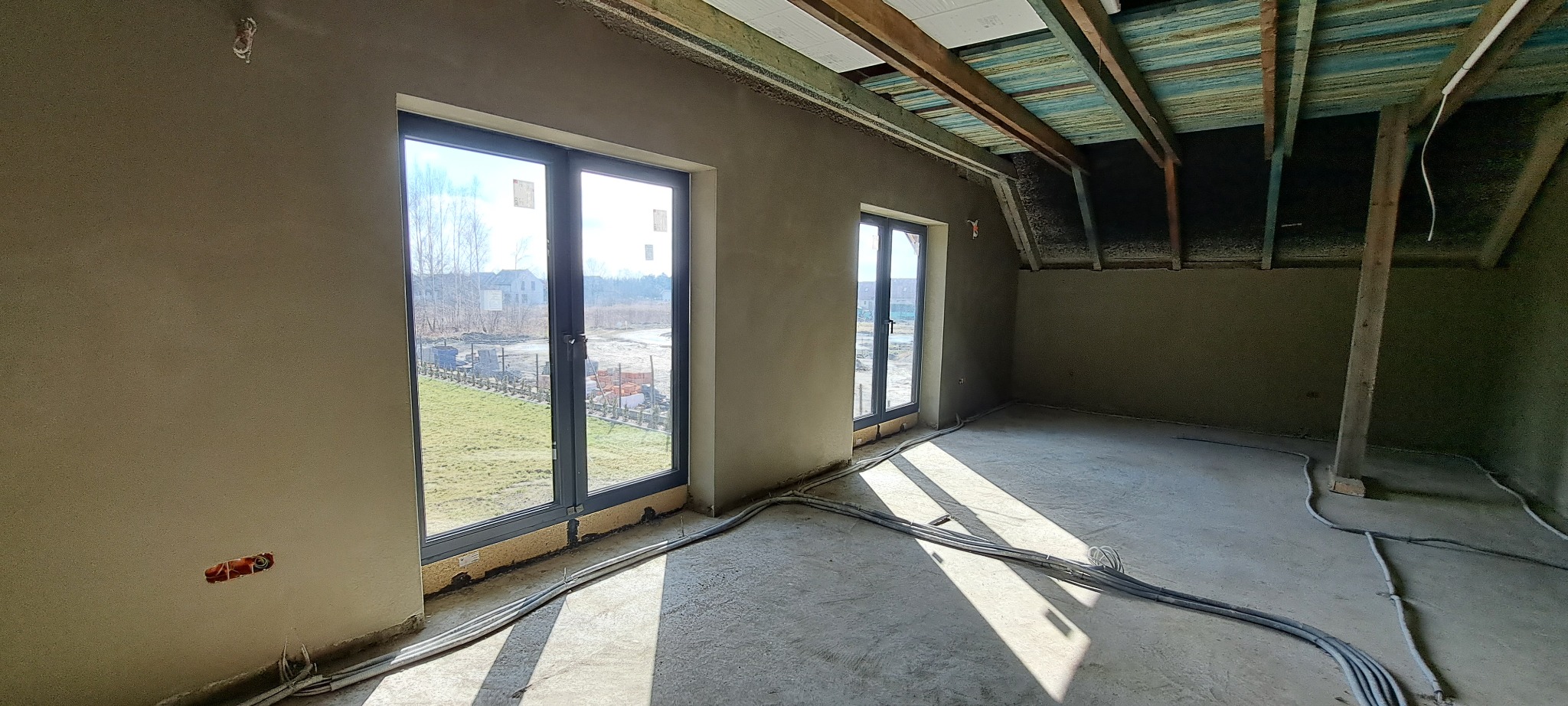Wnętrze domu w stanie surowym po nałożeniu tynków na ściany, widoczne okna z widokiem na zewnątrz, wiązki kabli elektrycznych na podłodze i surowy drewniany strop poddasza.