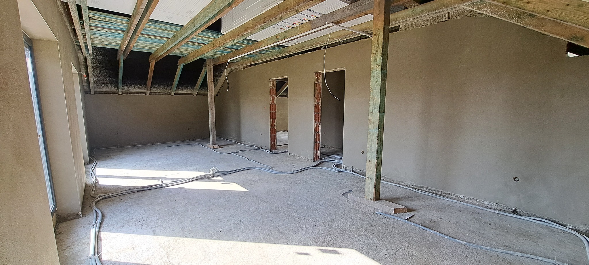 Surowe wnętrze po nałożeniu tynku maszynowego; widoczne drewniane elementy konstrukcyjne dachu, ściany w szarym tynku, instalacje elektryczne na podłodze i otwory drzwiowe w stanie surowym.