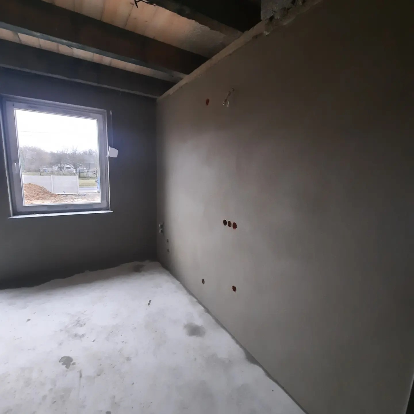 Wnętrze pokoju w trakcie prac wykończeniowych: ściany pokryte świeżym tynkiem, widoczne otwory na gniazdka elektryczne, surowy betonowy sufit i okno z widokiem na podwórze.