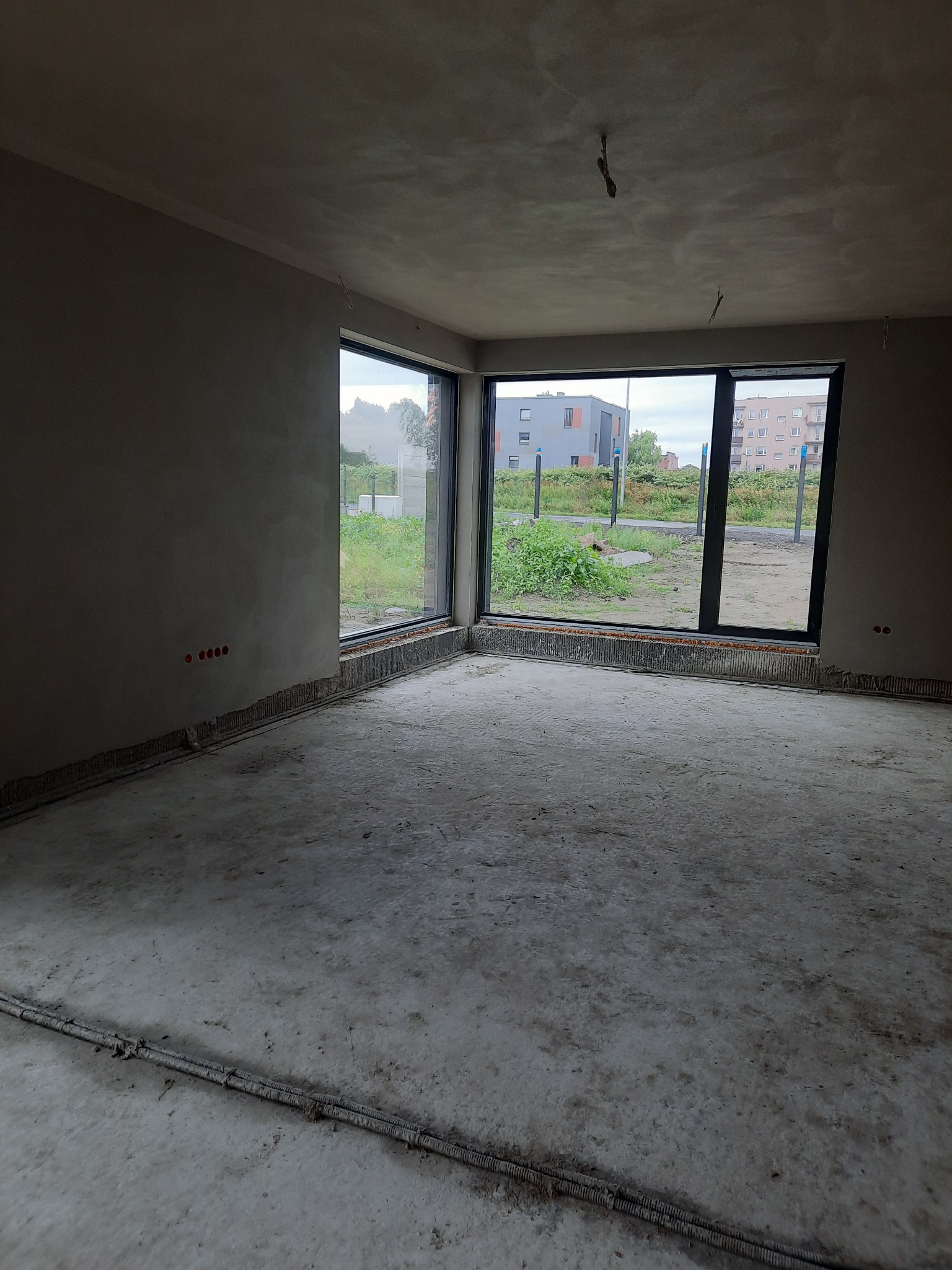 Surowe wnętrze z oknami, ściany pokryte świeżym tynkiem maszynowym, widoczne wystające kable elektryczne, betonowa podłoga w trakcie prac wykończeniowych.