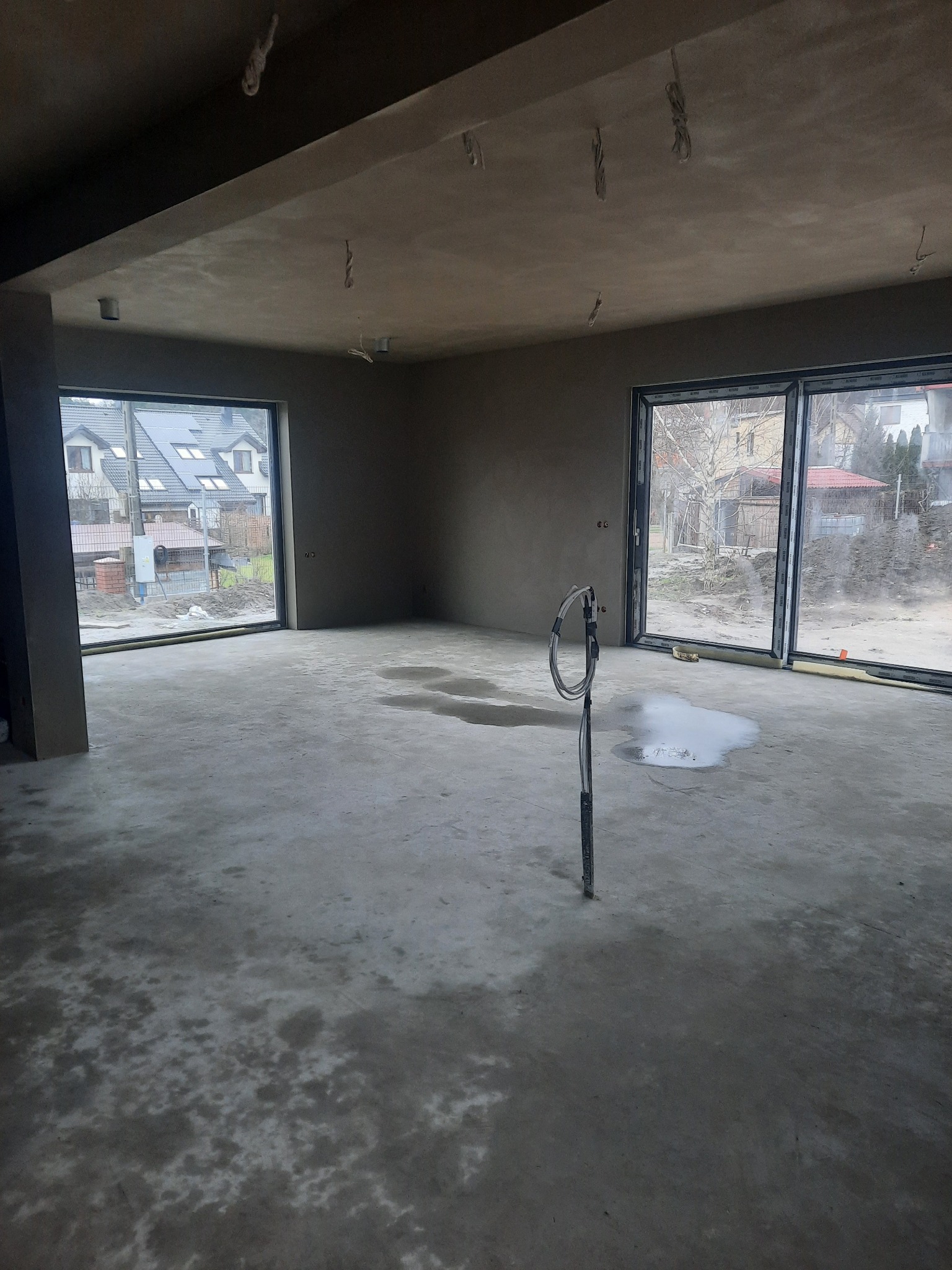 Wnętrze surowego domu z nałożonymi tynkami maszynowymi, widoczne ślady wilgoci na posadzce i wystające przewody elektryczne z sufitu, duże okna z widokiem na zabudowę jednorodzinną.
