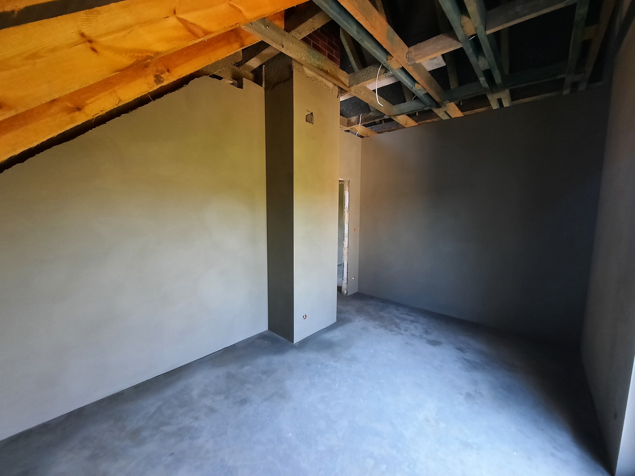 Wnętrze poddasza po nałożeniu gładzi gipsowej, widoczne drewniane elementy konstrukcji dachu, pionowy filar obłożony tynkiem oraz fragment muru z cegły w górnej części filara.