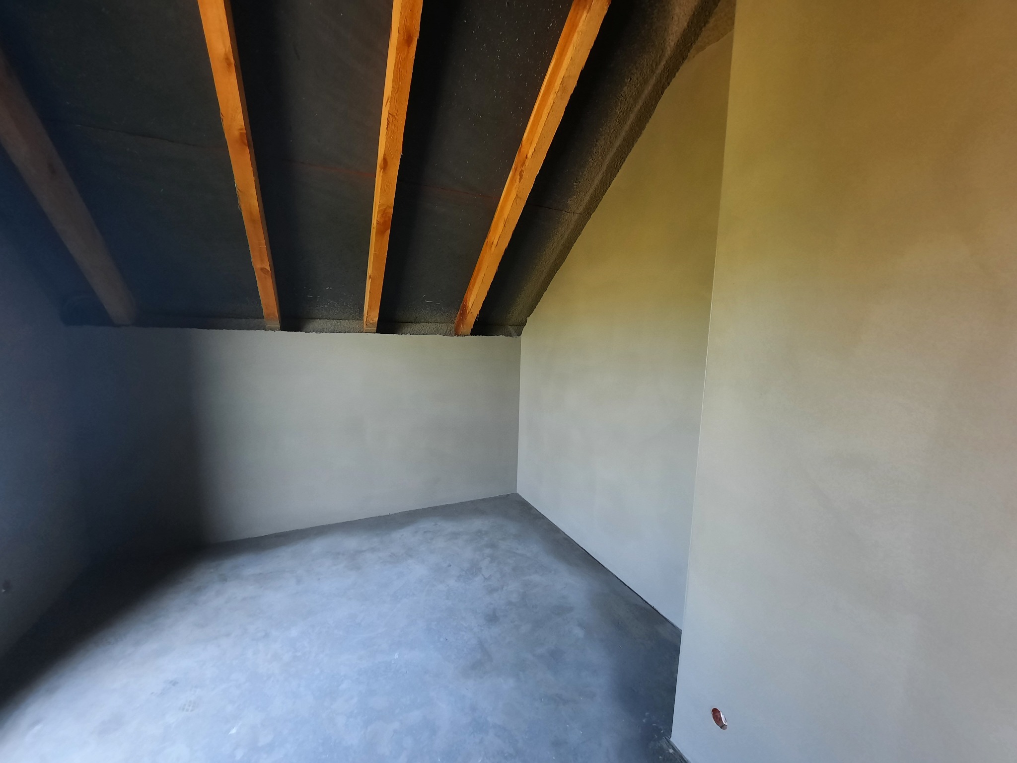 Surowe wnętrze poddasza po nałożeniu gładzi cementowej na ściany i podłogę, widoczne drewniane belki stropowe i czarna membrana dachowa.