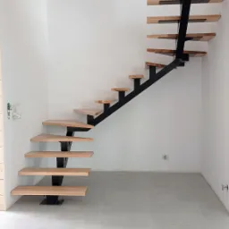 Nowoczesne schody wspornikowe z drewnianymi stopniami i czarną metalową konstrukcją w minimalistycznym wnętrzu z szarą podłogą i białymi ścianami.