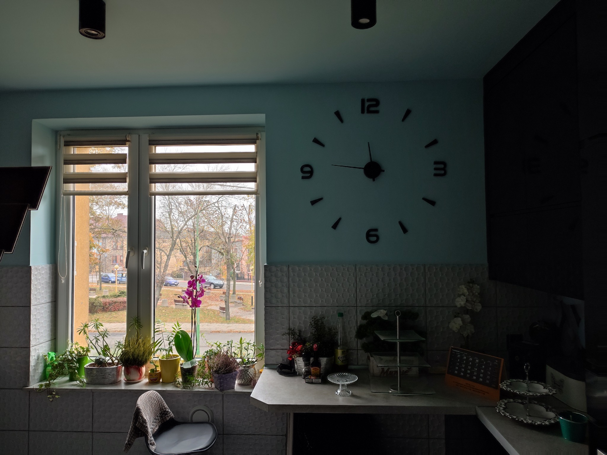 Jasne wnętrze kuchni z oknem, na parapecie rośliny doniczkowe, za oknem widok na ulicę, na ścianie duży zegar bez tarczy, na blacie akcesoria kuchenne i dekoracje.