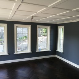 Usługi remontowo budowlane - Wnętrze pokoju po zakończonym malowaniu ścian na ciemny kolor, widoczne trzy okna z białymi ramami, biała listwa przypodłogowa, parkiet ułożony w jodełkę oraz dekoracyjny kaseton na suficie...
