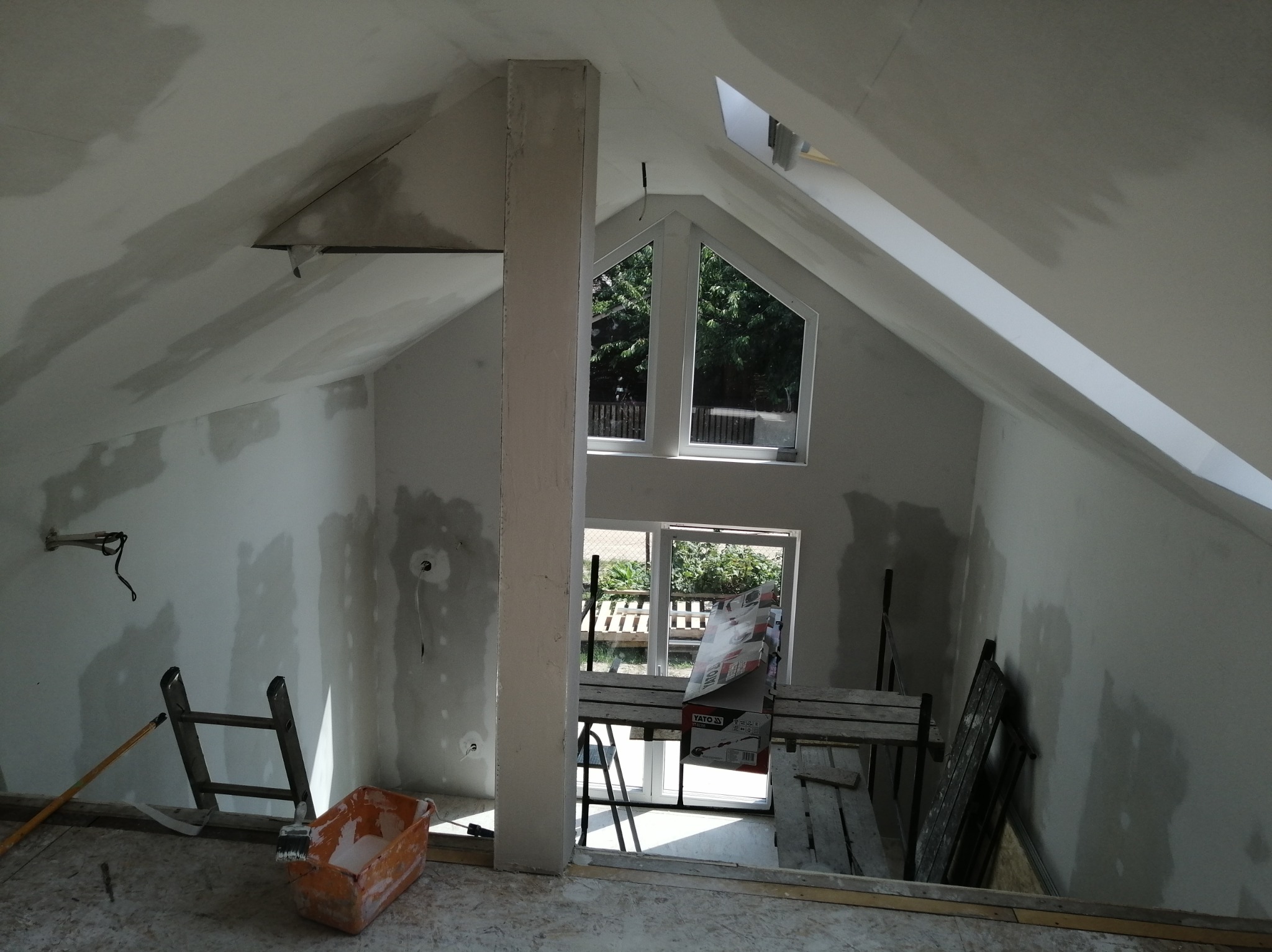 Poddasze w trakcie prac wykończeniowych, ściany i sufit zagruntowane, widoczne narzędzia malarskie, drabina i rusztowanie.