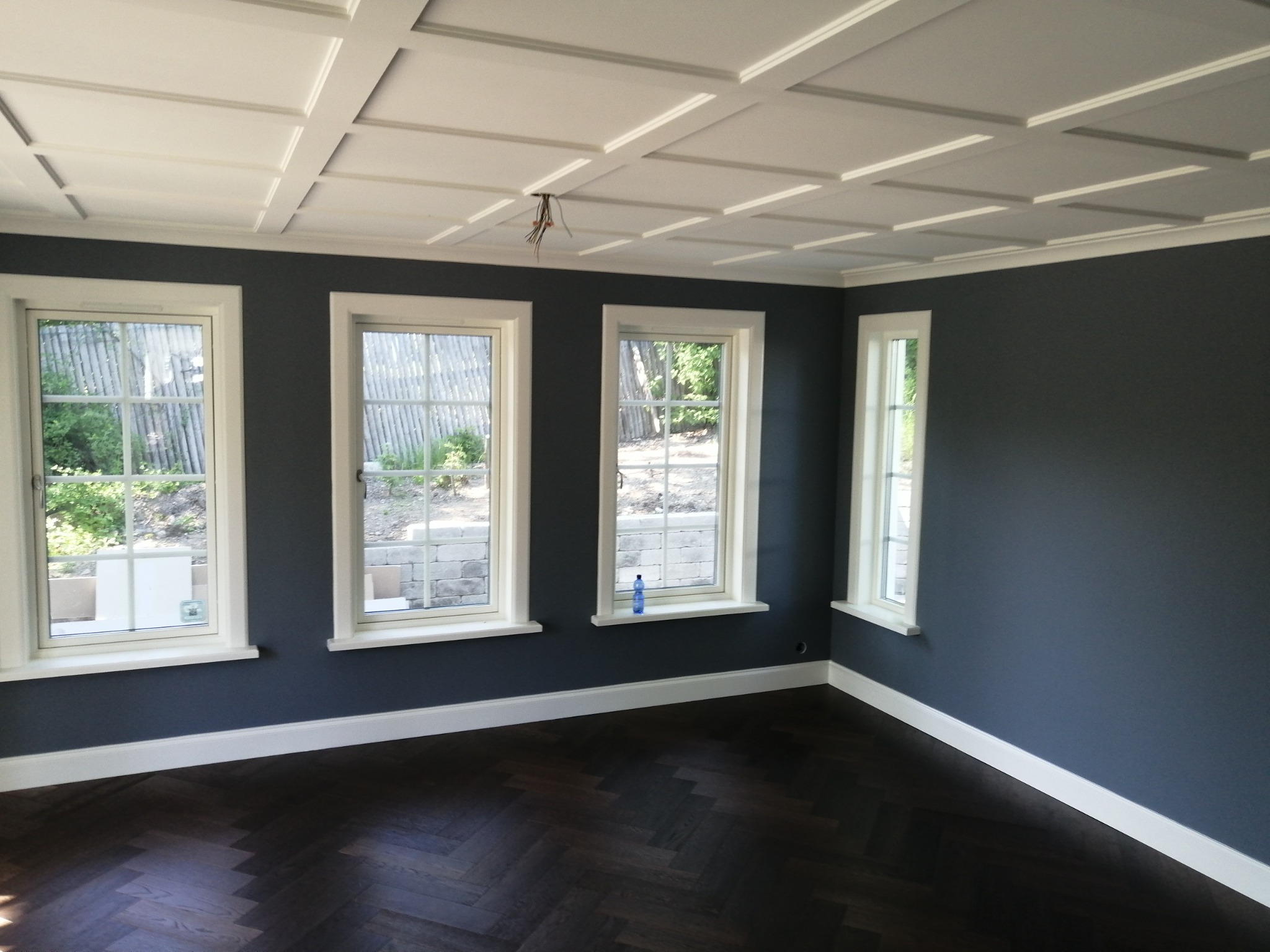 Wnętrze pokoju po zakończonym malowaniu ścian na ciemny kolor, widoczne trzy okna z białymi ramami, biała listwa przypodłogowa, parkiet ułożony w jodełkę oraz dekoracyjny kaseton na suficie...