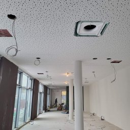 FIRMA HANDLOWO-USŁUGOWA AD-BUD ADAM MICKIEWICZ - Wnętrze w trakcie montażu oświetlenia: sufit podwieszany z otworami, wystające przewody, kolumny i okna. Widoczne ślady prac remontowych na podłodze.