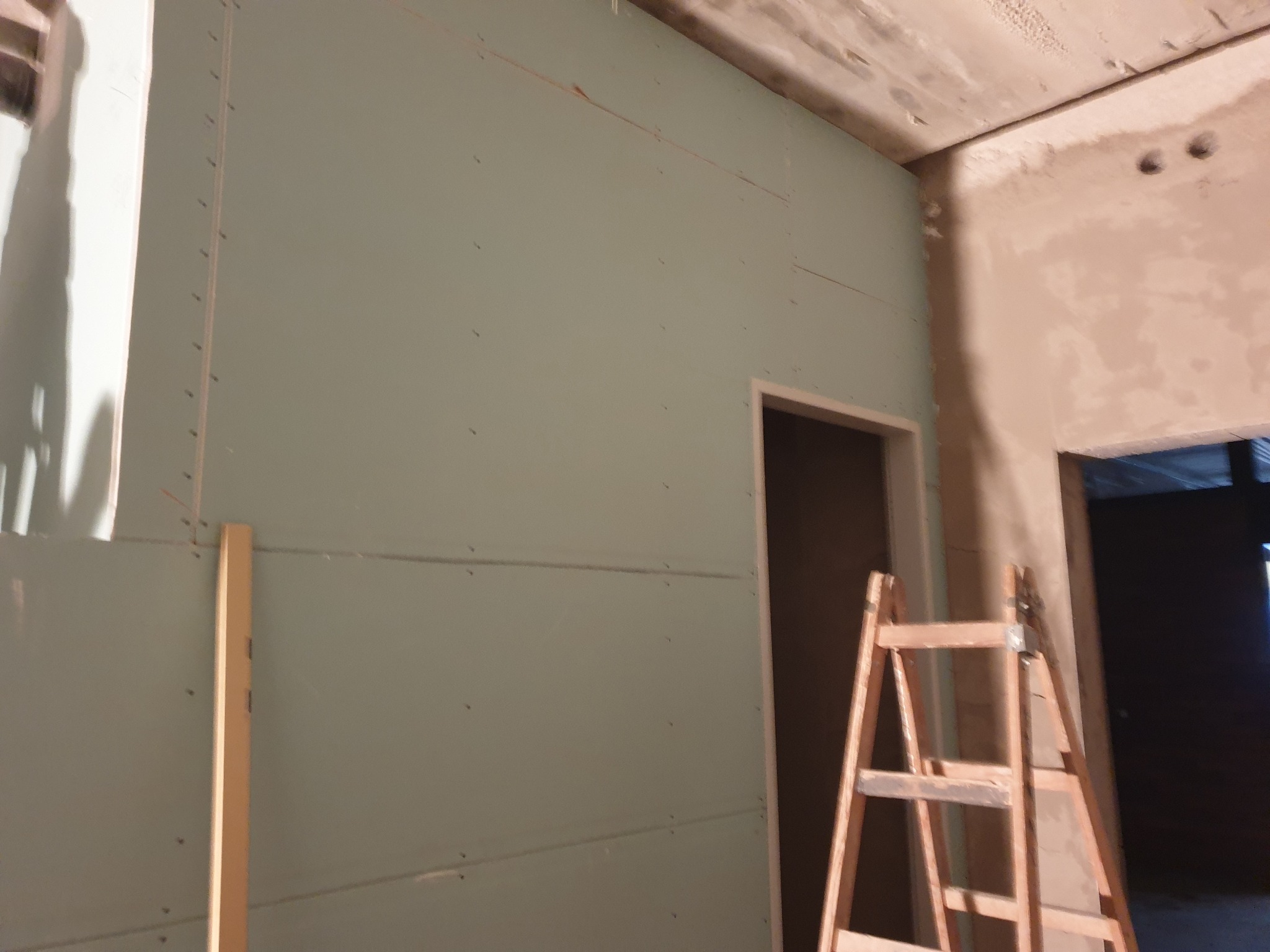 Ściana z płyt gipsowo-kartonowych w trakcie montażu, widoczna drabina i surowy strop. Etap prac wykończeniowych wewnątrz budynku.