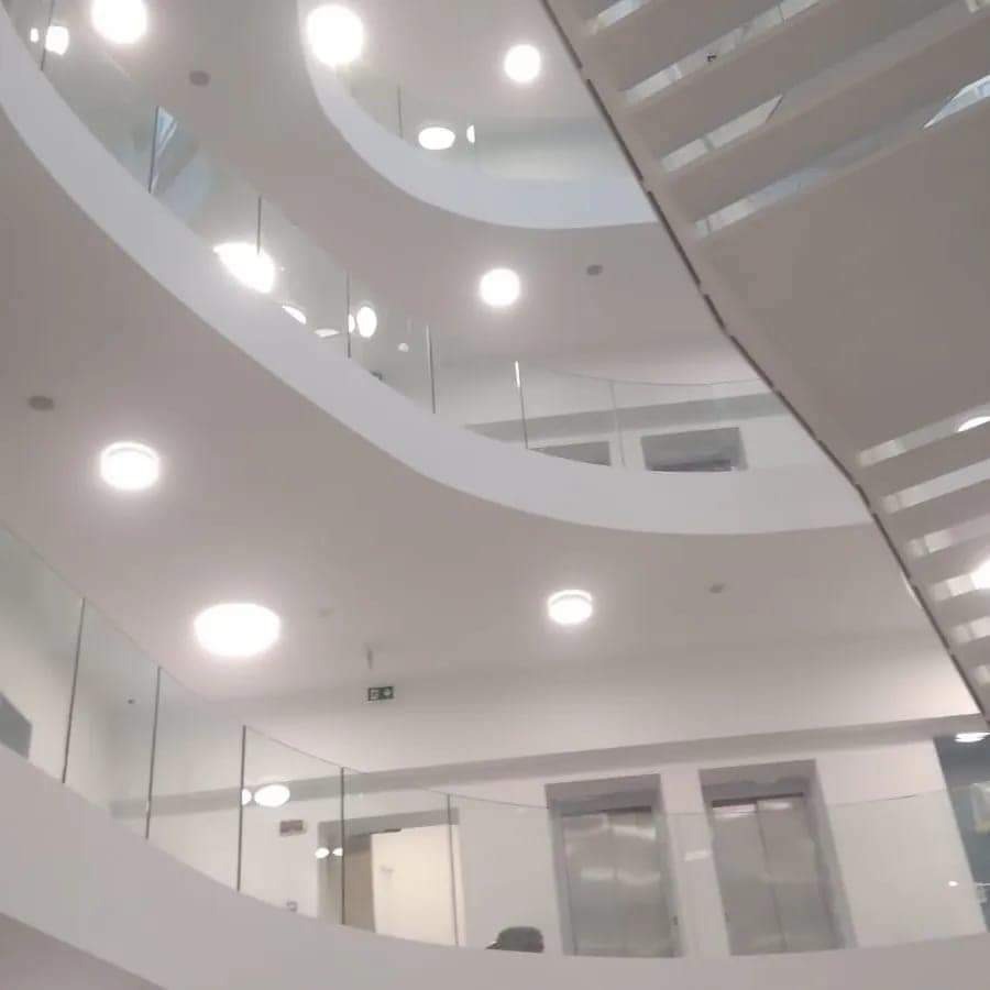 Nowoczesne wnętrze budynku z białymi, zakrzywionymi balkonami i szklanymi balustradami. Widoczne oświetlenie sufitowe i fragment schodów. Dwa srebrne szyby windy.