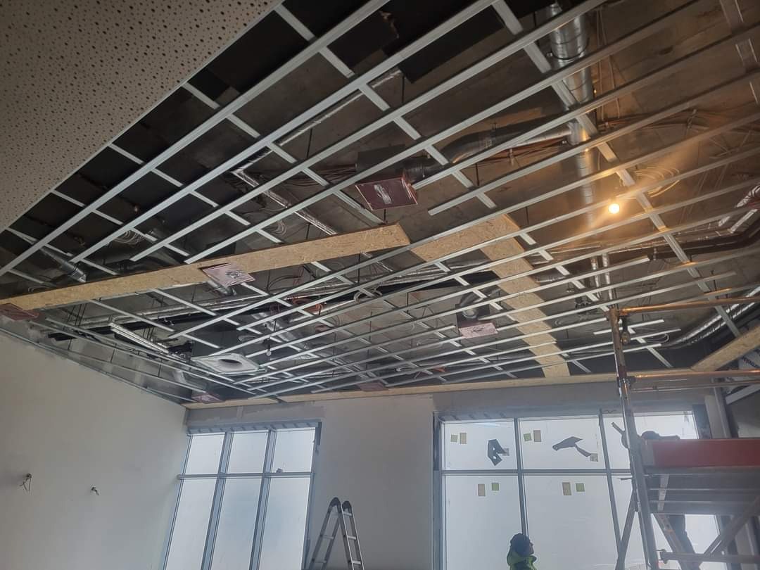 Montaż sufitu podwieszanego z widoczną konstrukcją metalową, instalacjami i częściowo zamontowanymi płytami. Widoczne okna i drabiny.