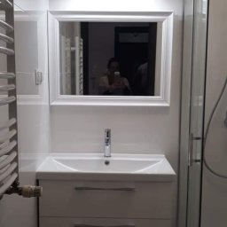 Zakład Remontowo Budowlany - Nowoczesna łazienka z białą umywalką nablatową, lustrem w białej ramie, grzejnikiem drabinkowym i kabiną prysznicową z zaokrąglonymi szybami. W lustrze odbija się osoba robiąca zdjęcie.