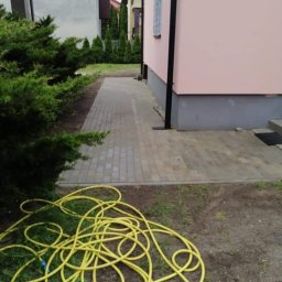 Zakład Remontowo Budowlany - Ułożona kostka brukowa tworząca ścieżkę obok domu z różową elewacją, żółty wąż ogrodowy leżący na ziemi, iglaki w tle.