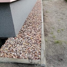 Zakład Remontowo Budowlany - Obsypanie kamieniami ozdobnymi fundamentów domu z widocznym fragmentem elewacji i chodnikiem z kostki, osoba grabiąca ziemię w tle.