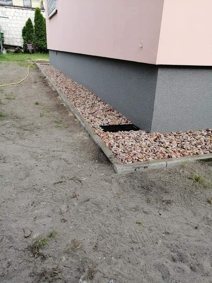 Wykończenie elewacji budynku z różową ścianą, szarą podmurówką i kamiennym drenażem wzdłuż fundamentów, widoczne ślady opon na ziemi.