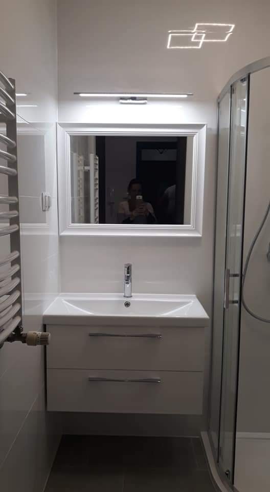 Nowoczesna łazienka z białą umywalką nablatową, lustrem w białej ramie, grzejnikiem drabinkowym i kabiną prysznicową z zaokrąglonymi szybami. W lustrze odbija się osoba robiąca zdjęcie.