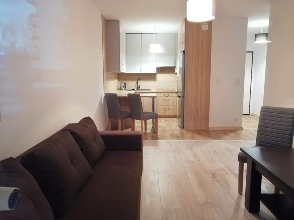 Jasne wnętrze mieszkania z drewnianą podłogą, brązową sofą, stołem z krzesłem i aneksem kuchennym z białymi szafkami i drewnianymi blatami, oświetlone wiszącymi lampami.