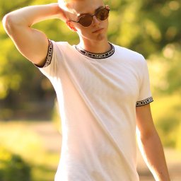 Dawid Morris - Młody mężczyzna z rudymi włosami w okularach przeciwsłonecznych, ubrany w białą koszulkę z napisami na kołnierzyku i rękawach, pozuje na tle zieleni w słoneczny dzień.