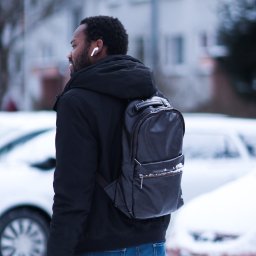 Dawid Morris - Mężczyzna z plecakiem idzie zimą, widoczny z profilu, w słuchawkach bezprzewodowych, na tle zaśnieżonych samochodów i budynku.