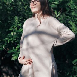 Dawid Morris - Kobieta w okularach, ubrana w beżowy komplet, trzyma dłonie na brzuchu na tle zielonych liści.