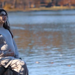 Dawid Morris - Kobieta w ciąży, ubrana w beżowy dres, siedzi na murku nad jeziorem, trzymając ręce na brzuchu i patrząc w dal.
