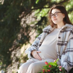 Dawid Morris - Uśmiechnięta kobieta w ciąży w okularach siedzi na ławce w parku, trzyma rękę na brzuchu, obok leżą tulipany.