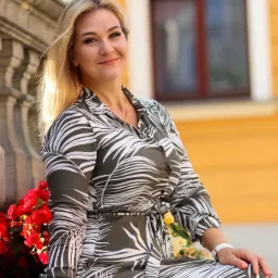 Uśmiechnięta blondynka w sukience w liście siedzi na kamiennym murku z czerwonymi kwiatami, w tle żółta fasada budynku z oknem.