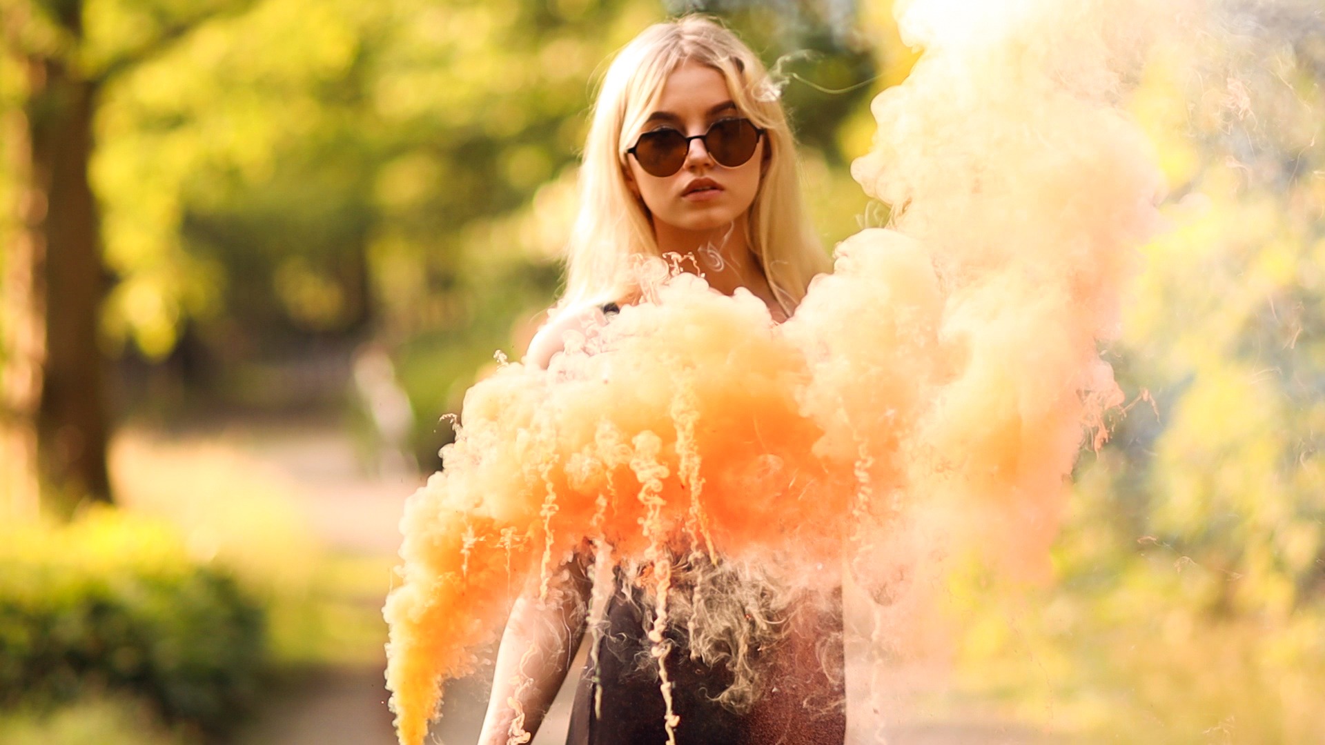 Blondwłosa kobieta w okularach przeciwsłonecznych pozuje z pomarańczowym dymem na tle rozmytej zieleni.