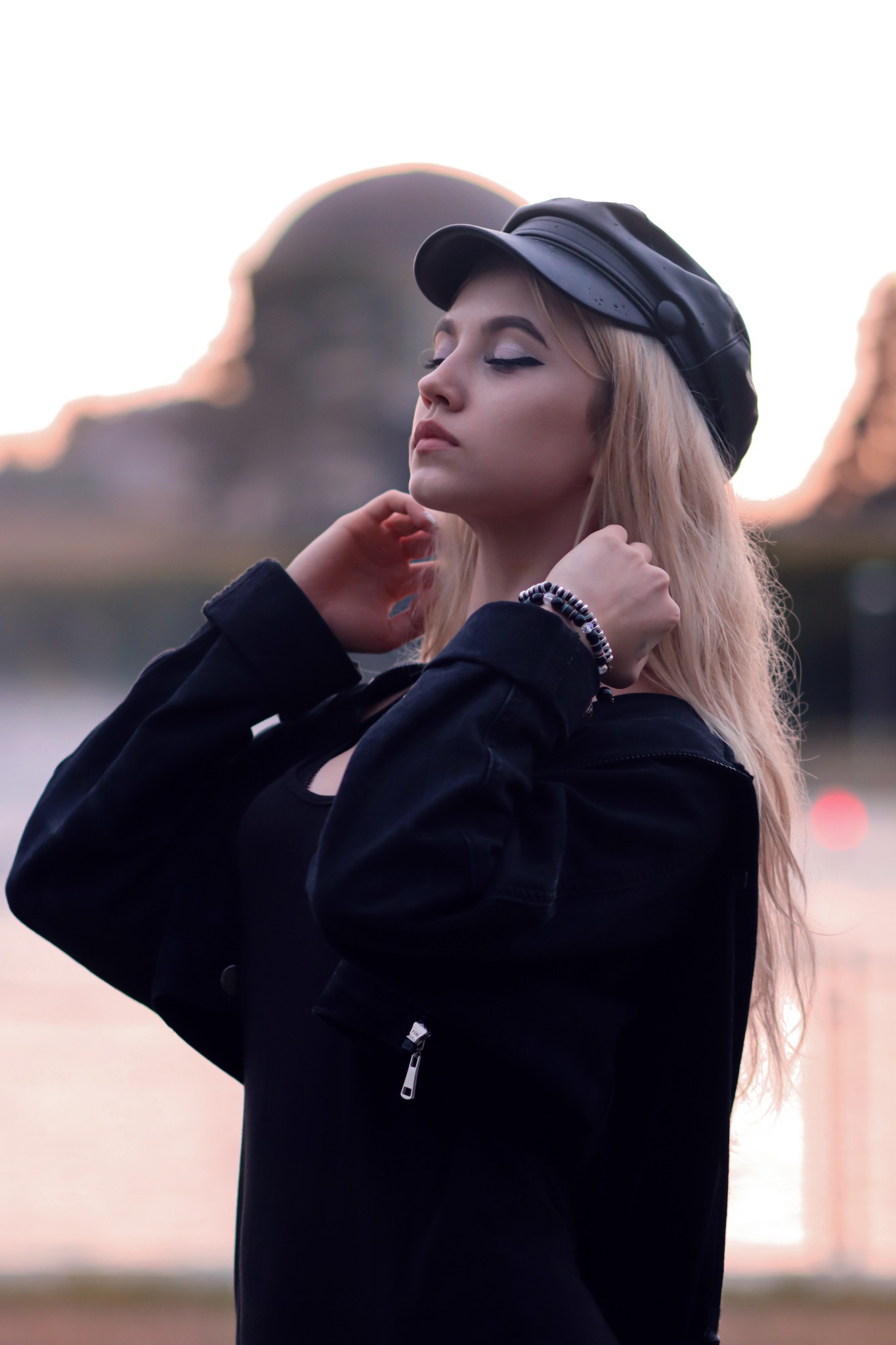 Portret młodej kobiety z blond włosami, w czarnej kurtce i czapce, z zamkniętymi oczami, na rozmytym tle miejskim o zmierzchu.