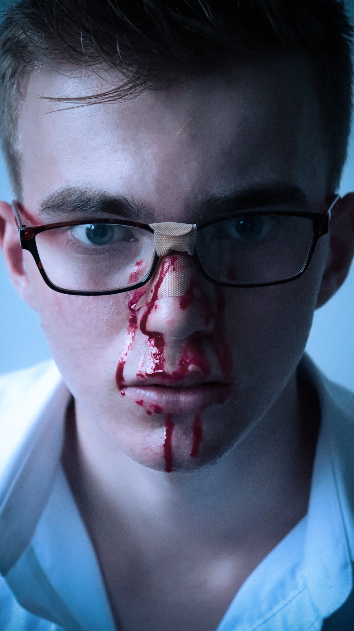 Portret młodego mężczyzny we Wrocławiu z krwawiącym nosem i plastrami, noszącego okulary i białą koszulę, w chłodnym oświetleniu.