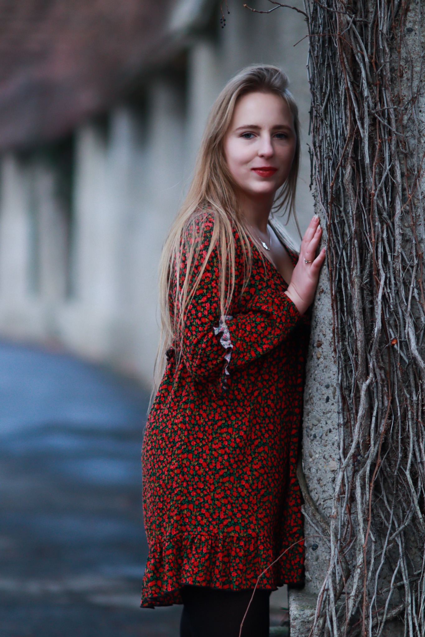 Blondwłosa kobieta w kwiecistej sukience opiera się o kamienny filar porośnięty suchymi pnączami, na tle rozmytej ulicy.