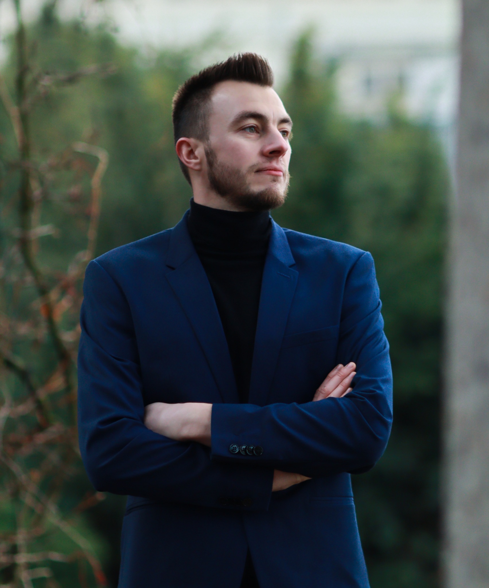 Młody mężczyzna w granatowym garniturze i czarnym golfie, z brodą i starannie ułożonymi włosami, stoi na tle zieleni z założonymi rękami i patrzy w dal.