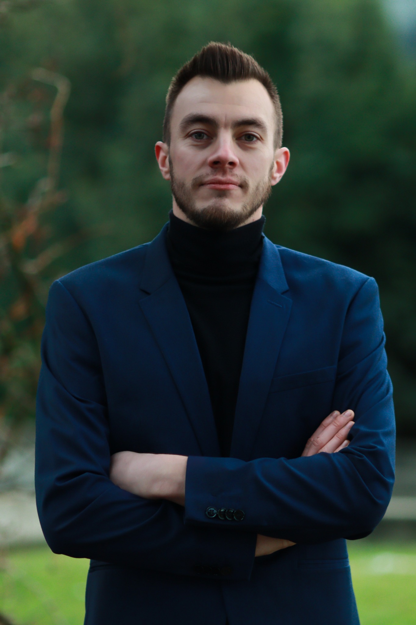 Portret młodego mężczyzny w ciemnoniebieskim garniturze i czarnym golfie, z założonymi rękami na tle zieleni.