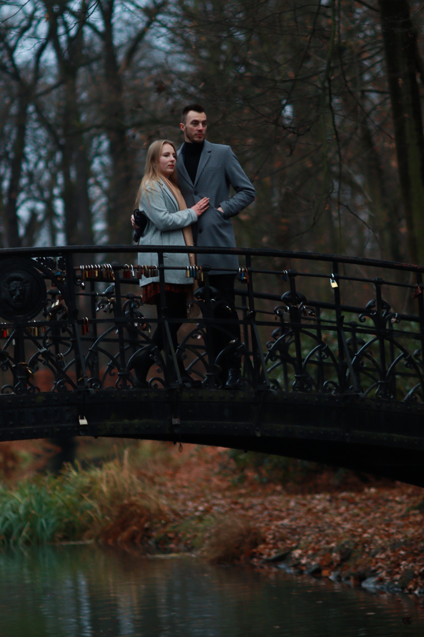 Para w eleganckich płaszczach stoi na moście ozdobionym kłódkami w jesiennym parku.