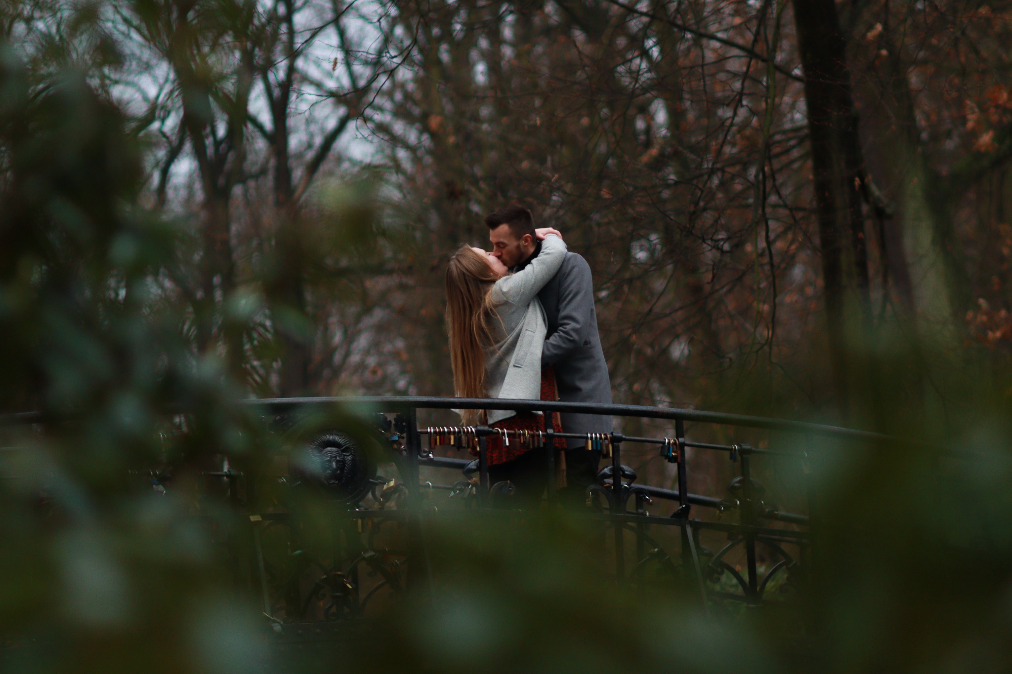 Zakochana para całuje się na moście ozdobionym kłódkami miłości, w tle drzewa bez liści, ujęcie przez rozmyte gałęzie na pierwszym planie.