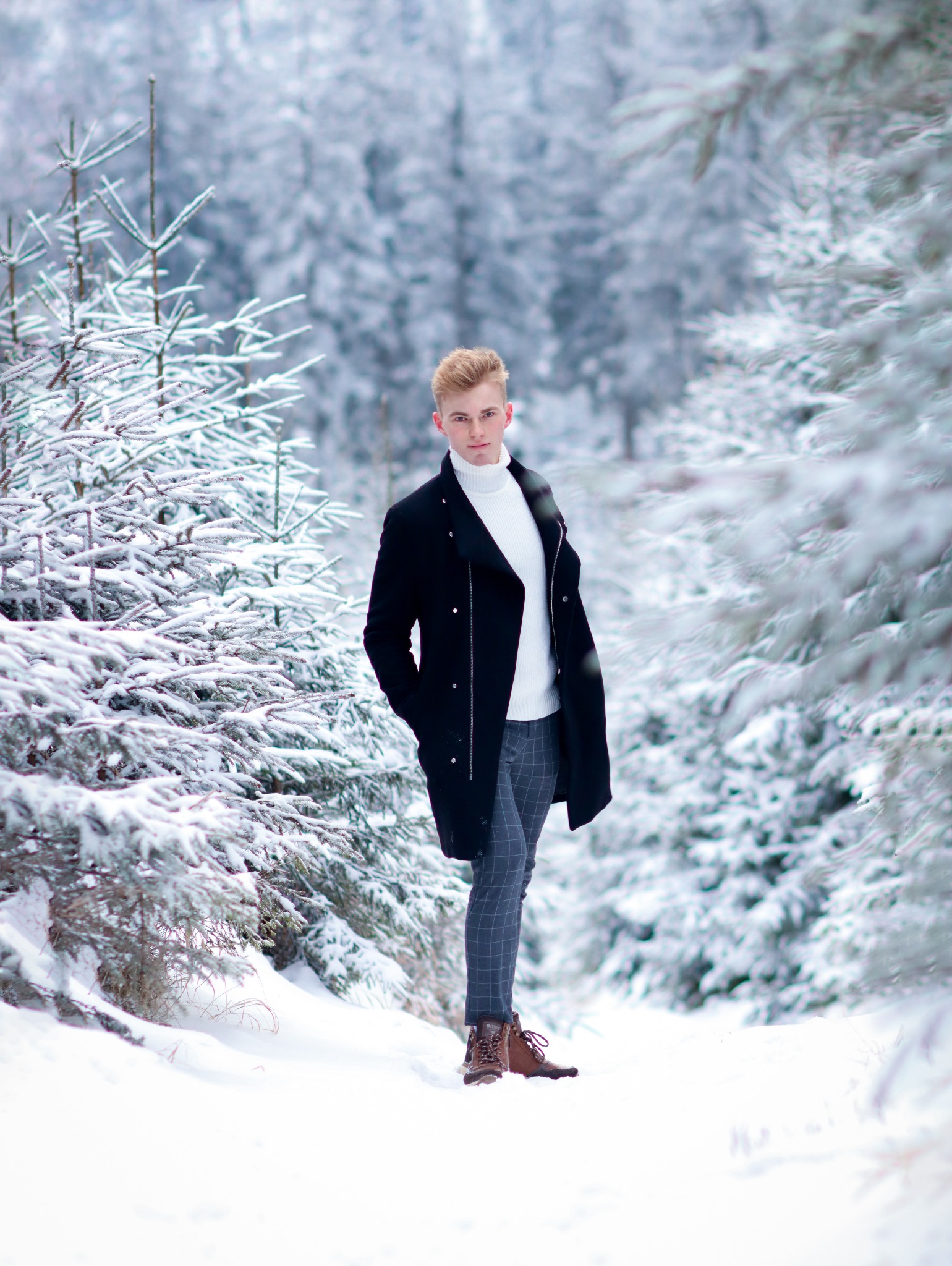 Młody mężczyzna w czarnym płaszczu i białym golfie, pozuje w zasypanym śniegiem lesie iglastym, z rękami w kieszeniach, w tle drzewa pokryte szronem.