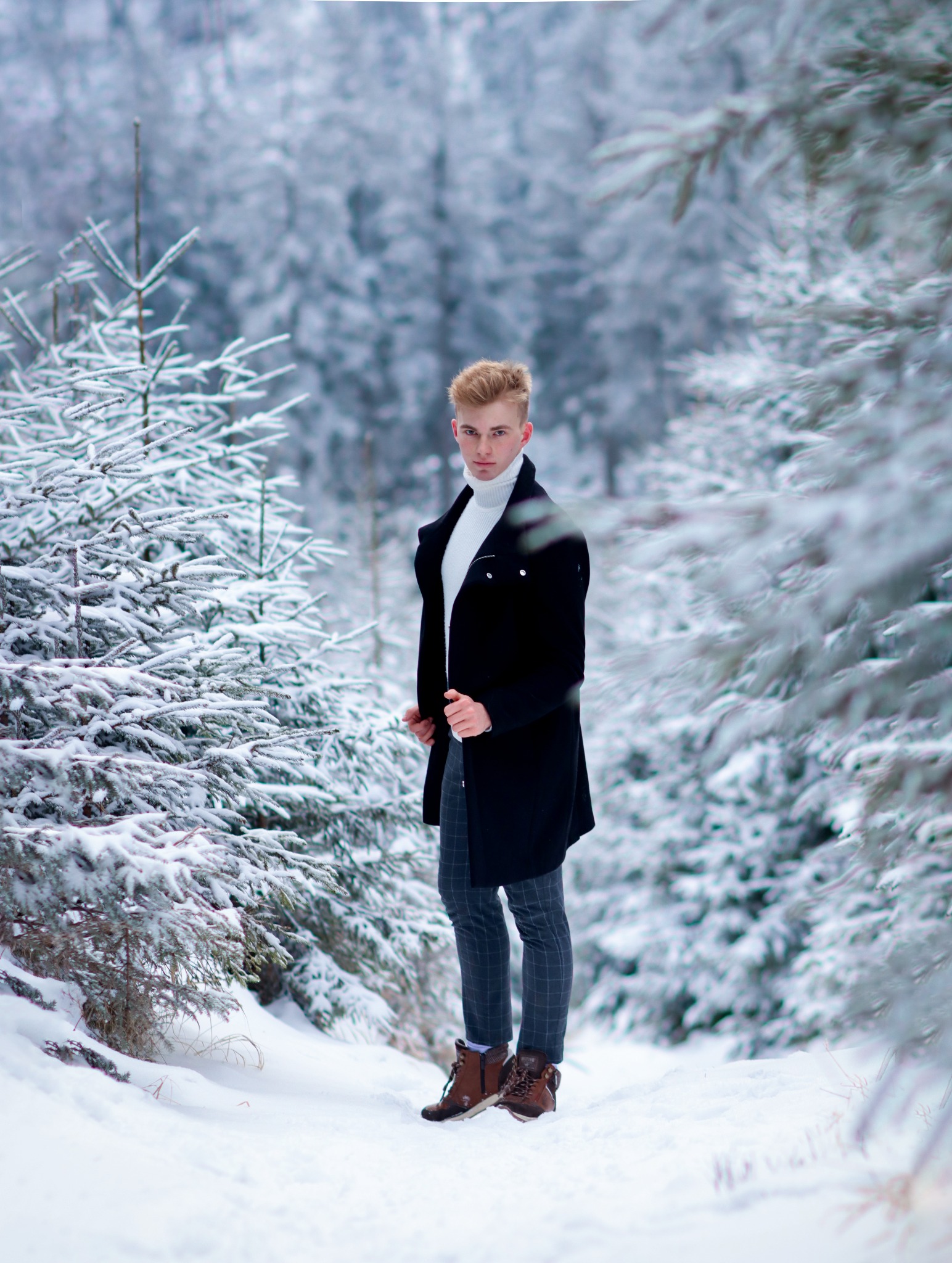Młody mężczyzna w czarnym płaszczu i białym golfie pozuje w zimowym lesie, otoczony ośnieżonymi drzewami.