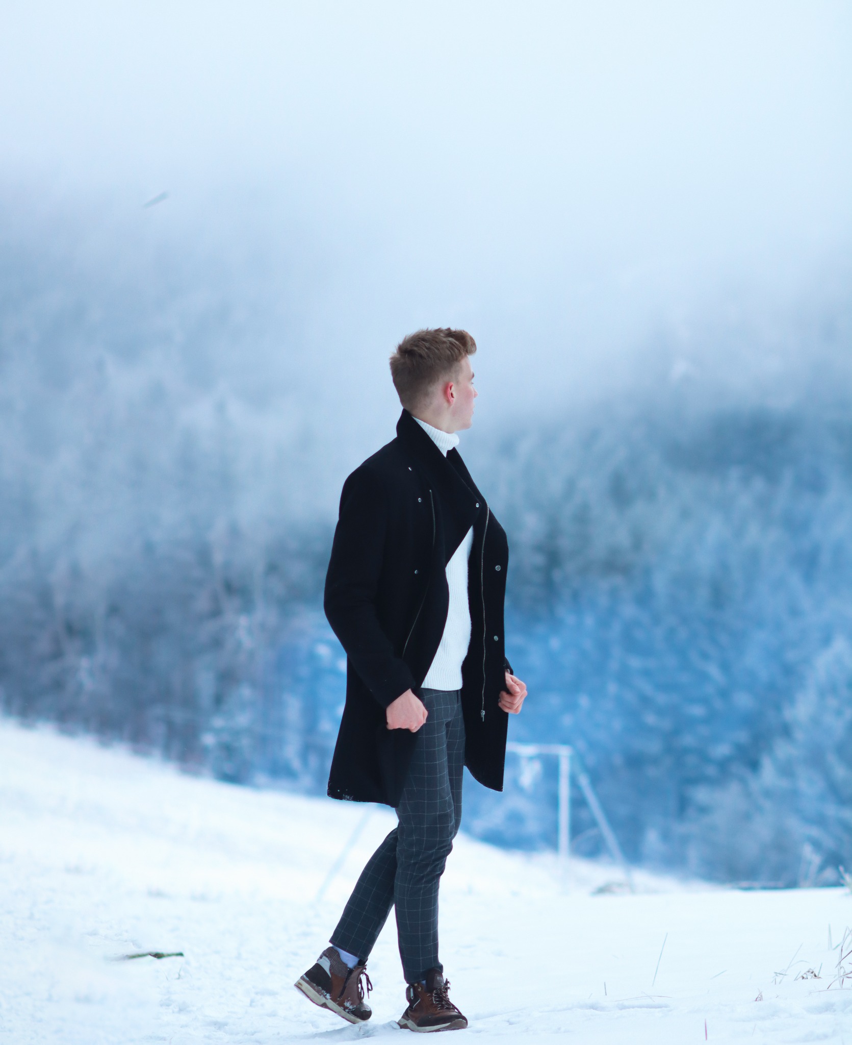 Mężczyzna w czarnym płaszczu, białym golfie i spodniach w kratę spaceruje po zasypanym śniegiem terenie z zalesionym tłem w zimowej scenerii.